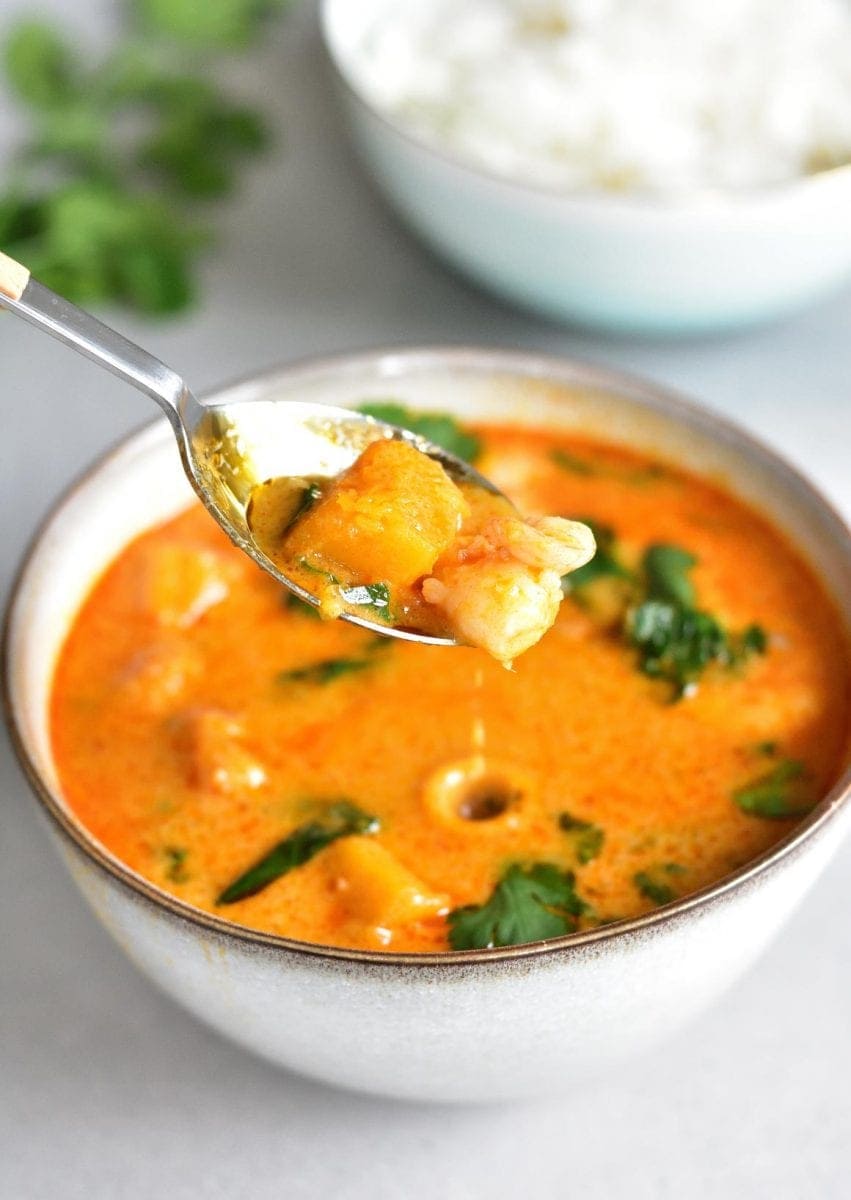 A spoon is scooping Thai red curry with shrimp and pumpkin from a bowl.