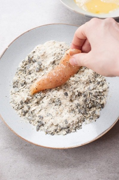 Chicken tenderloing is being coated in breadcrumbs and pumpkin seed mixture