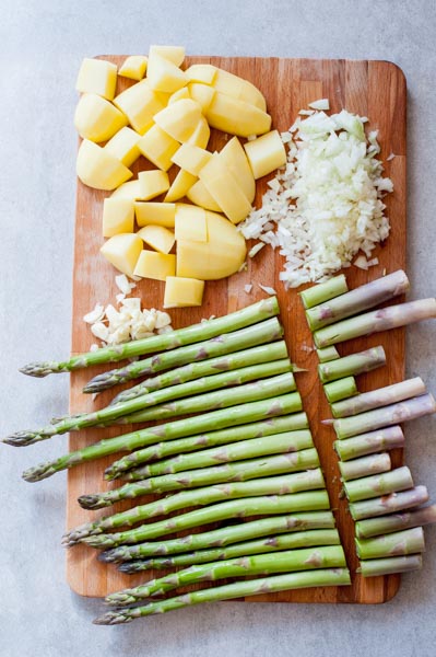 trimmed asparagus ends on a chopping board, chopped potatoes, onion and garlic