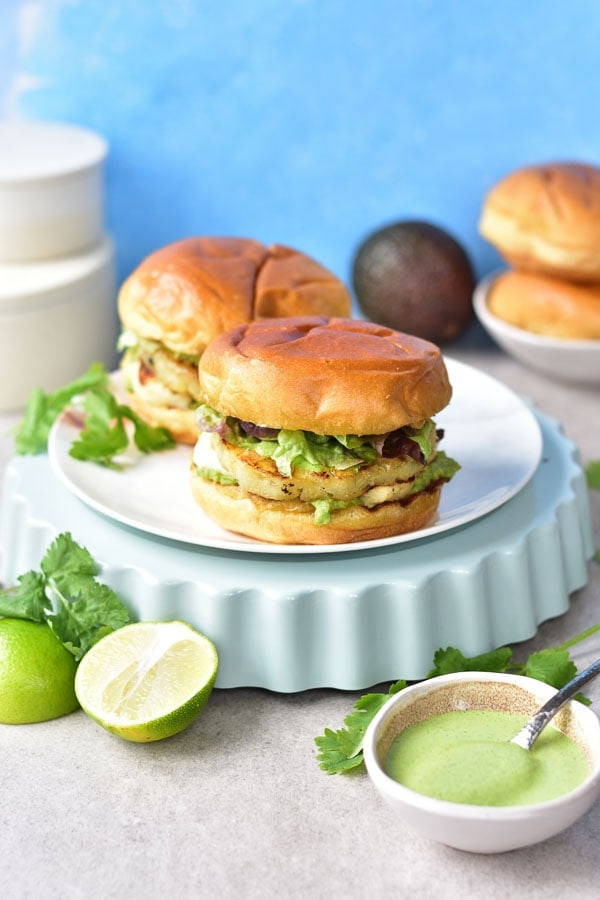 two halloumi burgers on a white plate, coriander, limes and avocado in the background