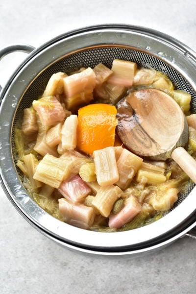 Cooked rhubarb on a fine mesh strainer, is being pressed with a wooden spoon