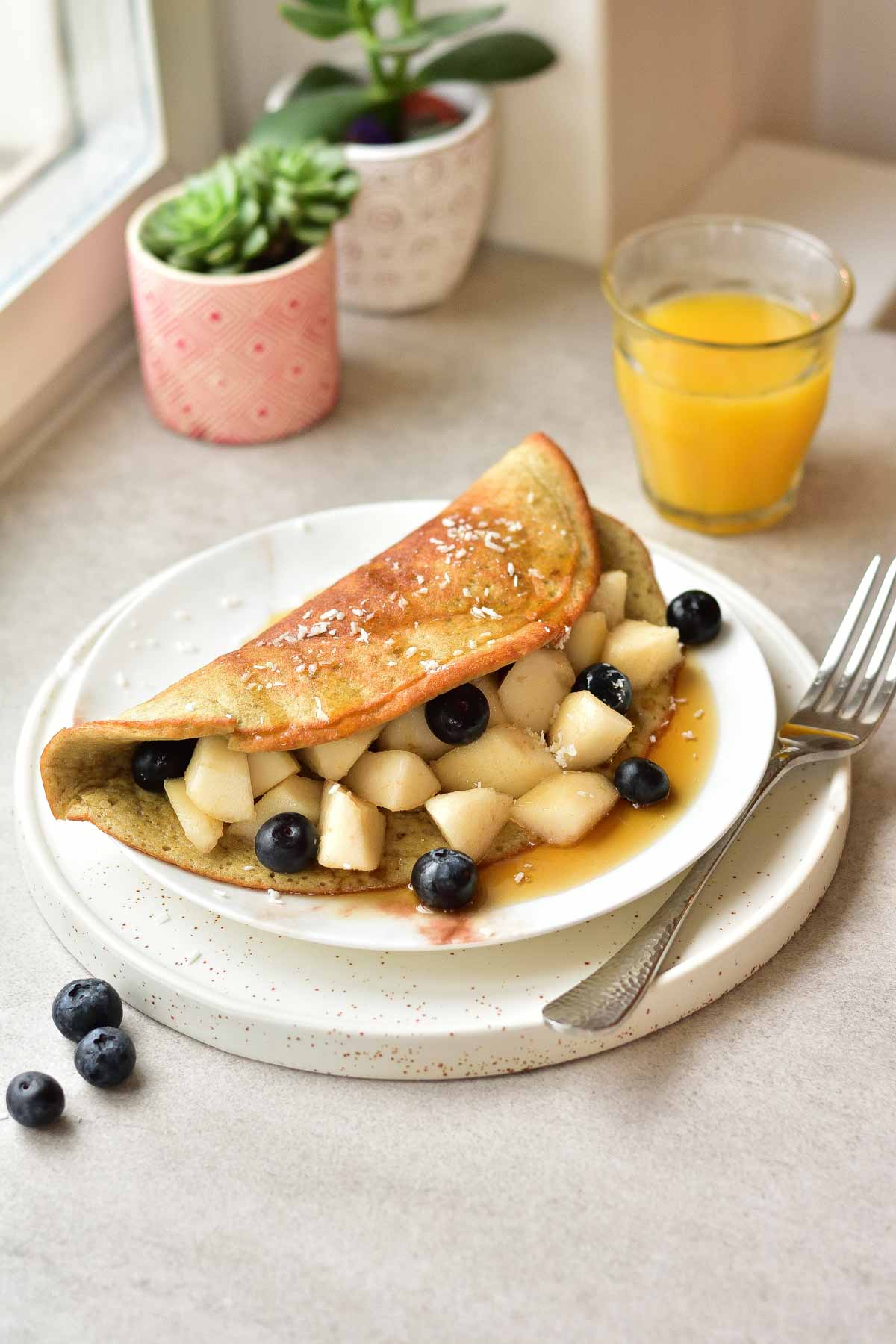 Banana omlette with pear and blueberries on a white plate, orange juice and plants in the background.