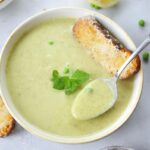 white plate with zucchini soup, spoon, mint leaves and cheese toast in the middle of the soup