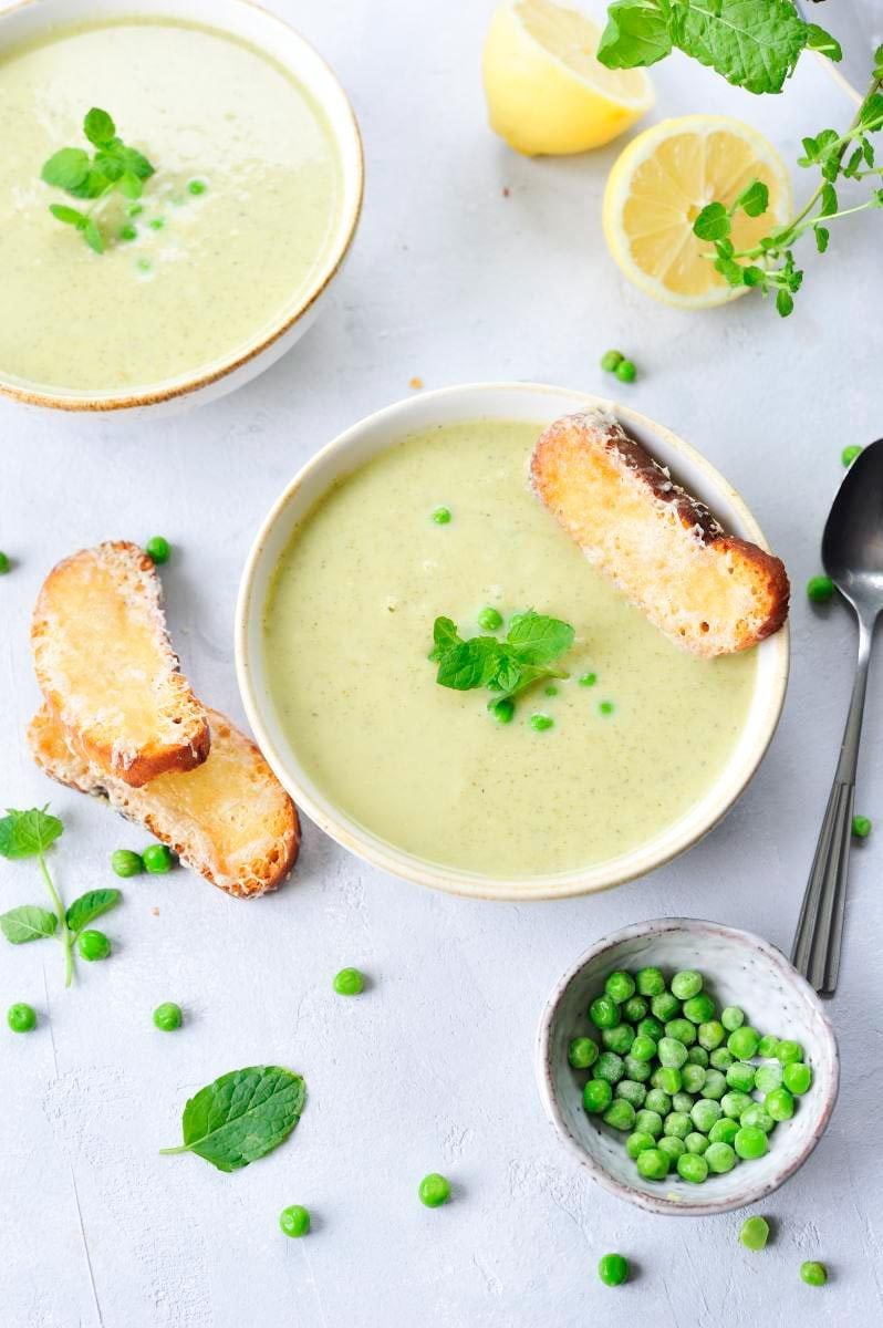 two white bowls with creamy zucchini soup, mint leaves, peas and cheese toasts are surrounding the bowls