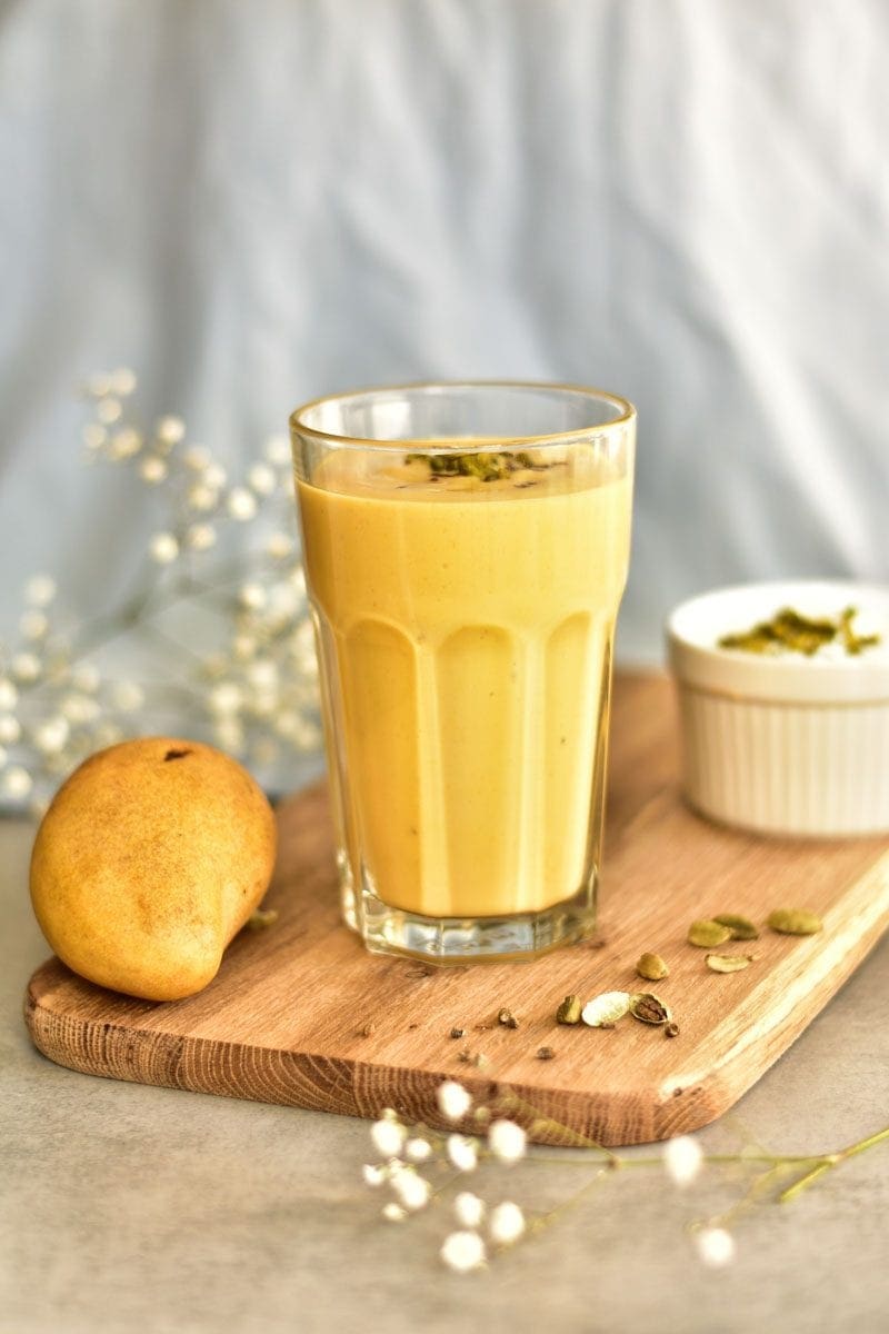 mango lassi w szklance, obok mango i nasiona kardamonu