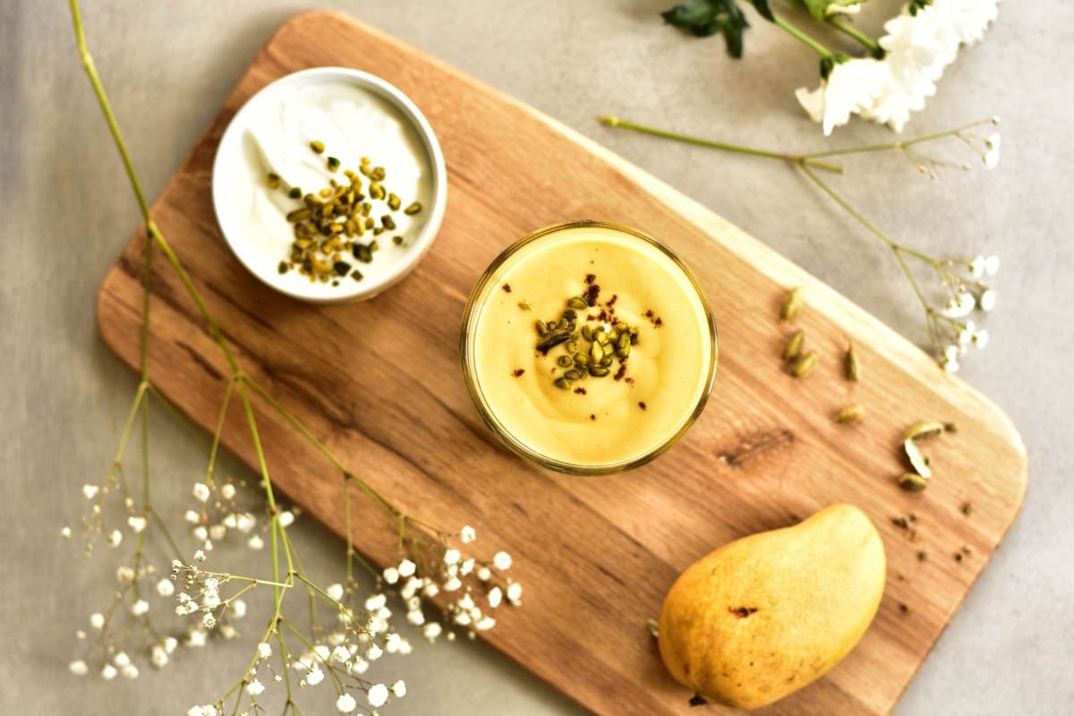 Overhead picture of mango lassi in a glass surrounded with yogurt, mango and flowers.