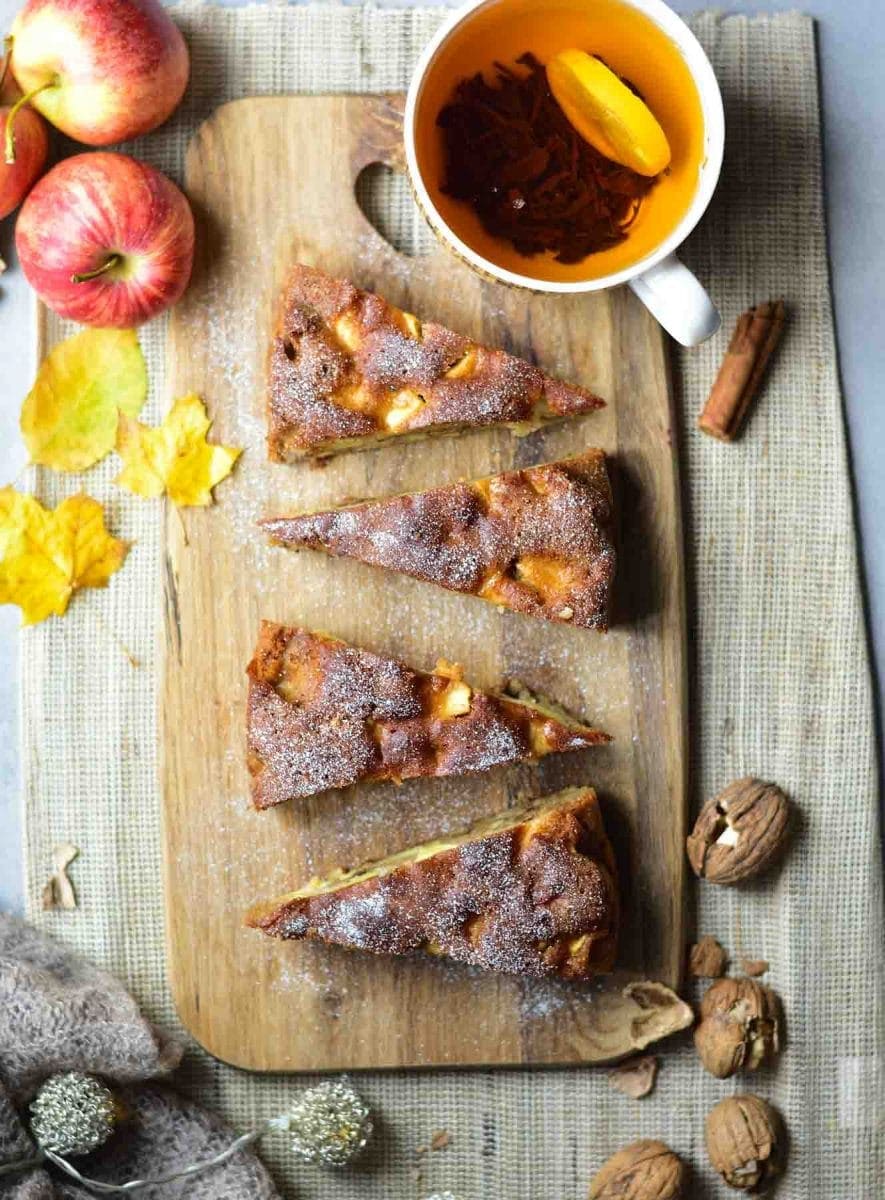 A couple of slices of apple walnut cake with honey on a wooden board. A cup of tea and apples in the background.