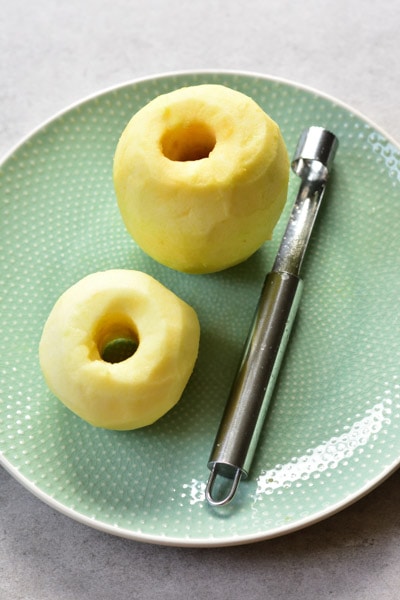 2 peeled and cored apples with an apple corer on a plate. Apple rings on a plate.
