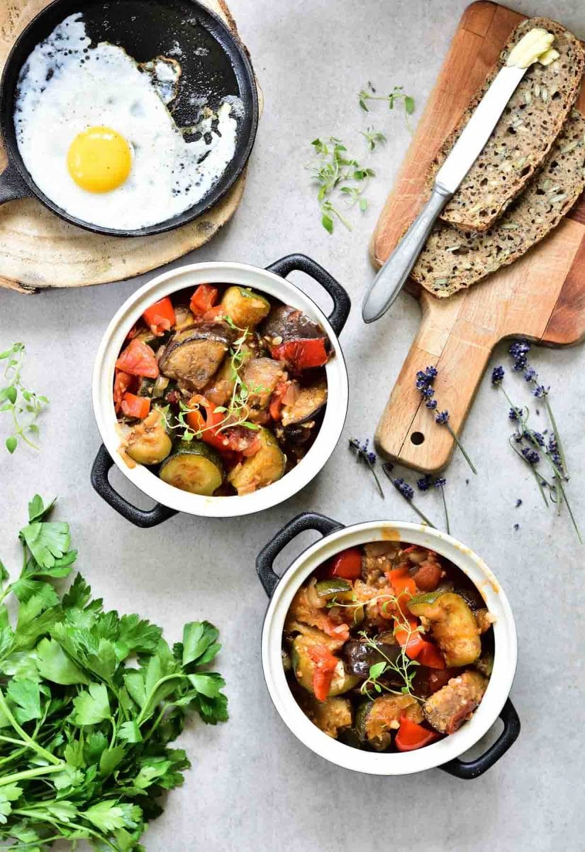 Two black pots with Ratatouille, fried egg in a pan, slices of bread with butter.