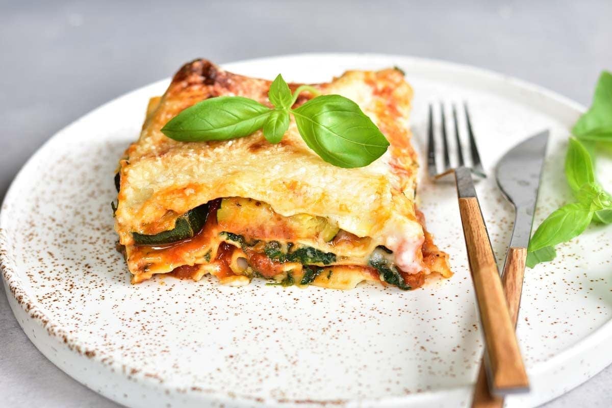 A serving of vegetarian lasagna with zucchini and spinach on a white plate, topped with basil leaves.