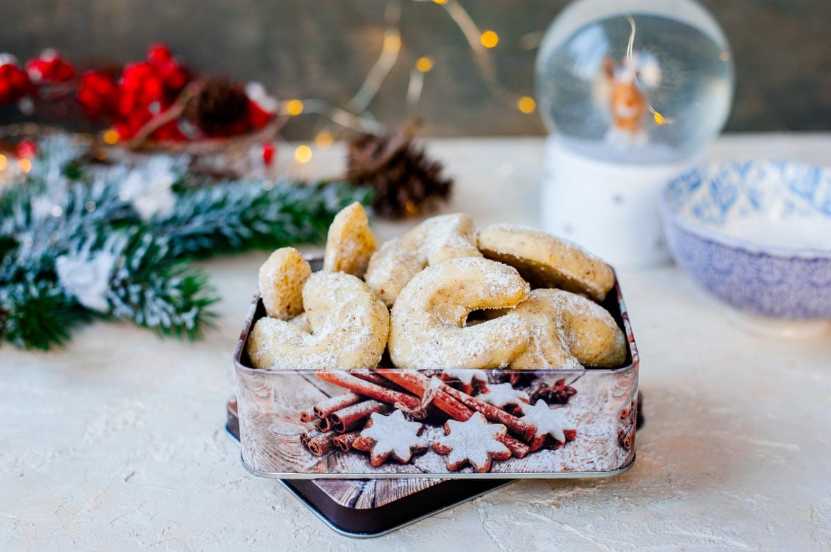 Vanilla crescent cookies in a cookie tin. Christmas lights and a tree branch in the background.