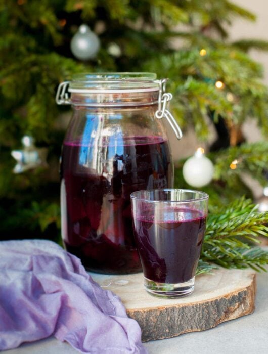 a big jar and a small glass filled with beet kvass, Christmass tree in the background