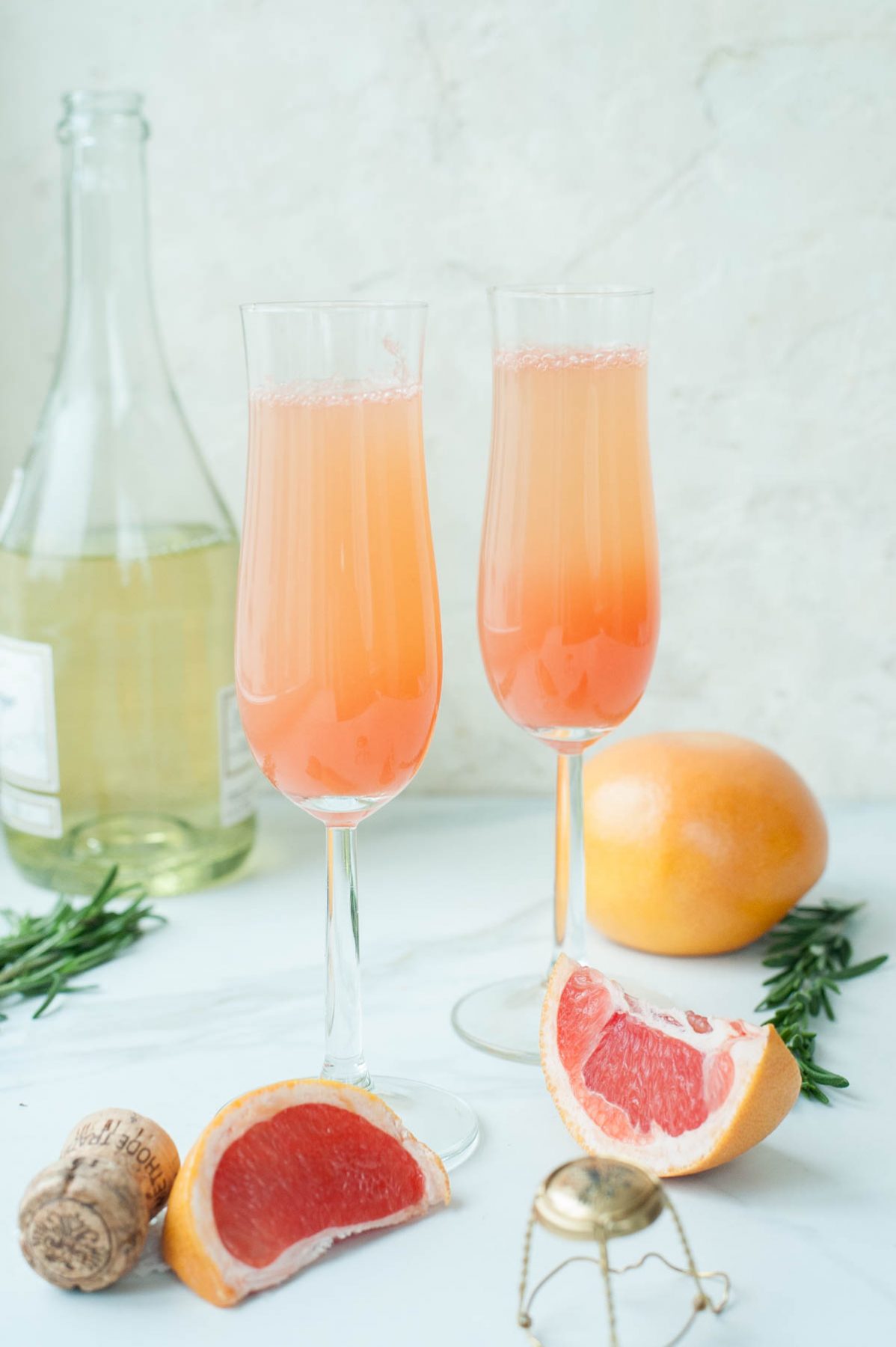 Two champagne glasses filled with grapefruit mimosa, grapefruit, rosemary and champagne in the background.