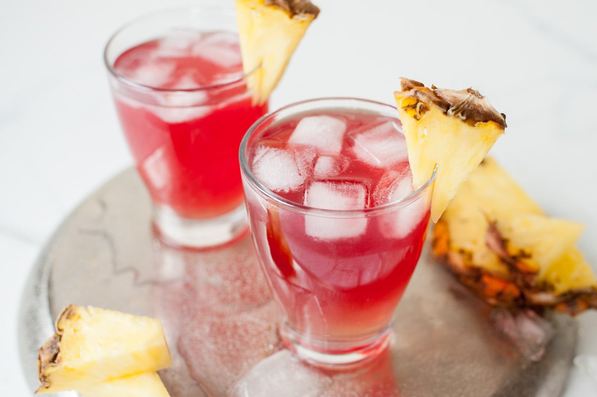 Two glasses with Malibu bay breeze drink on a silver tray garnished with pineapple wedges.