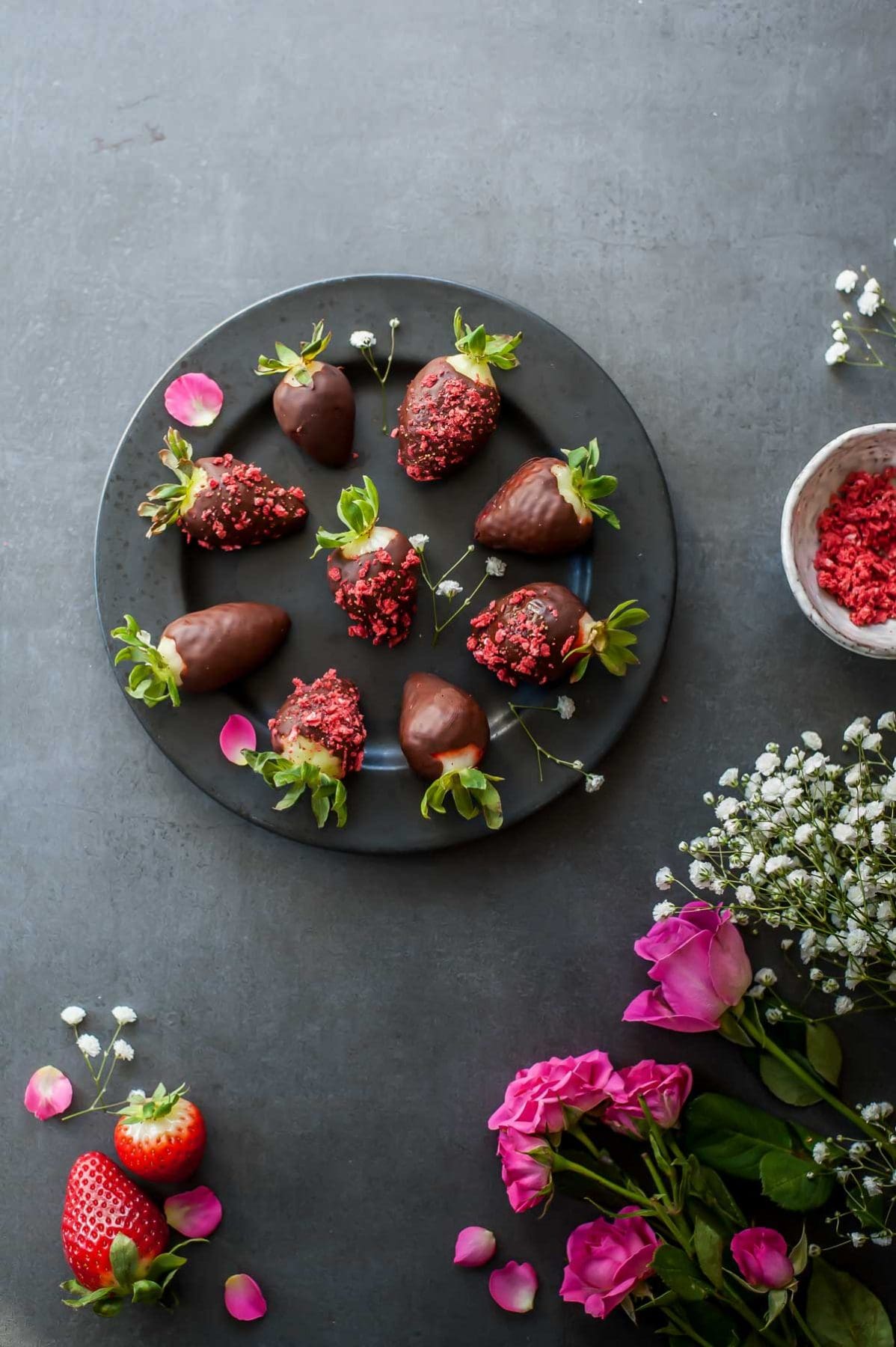 Chocolate covered strawberries on a black plate, roses and flowers around the plate.