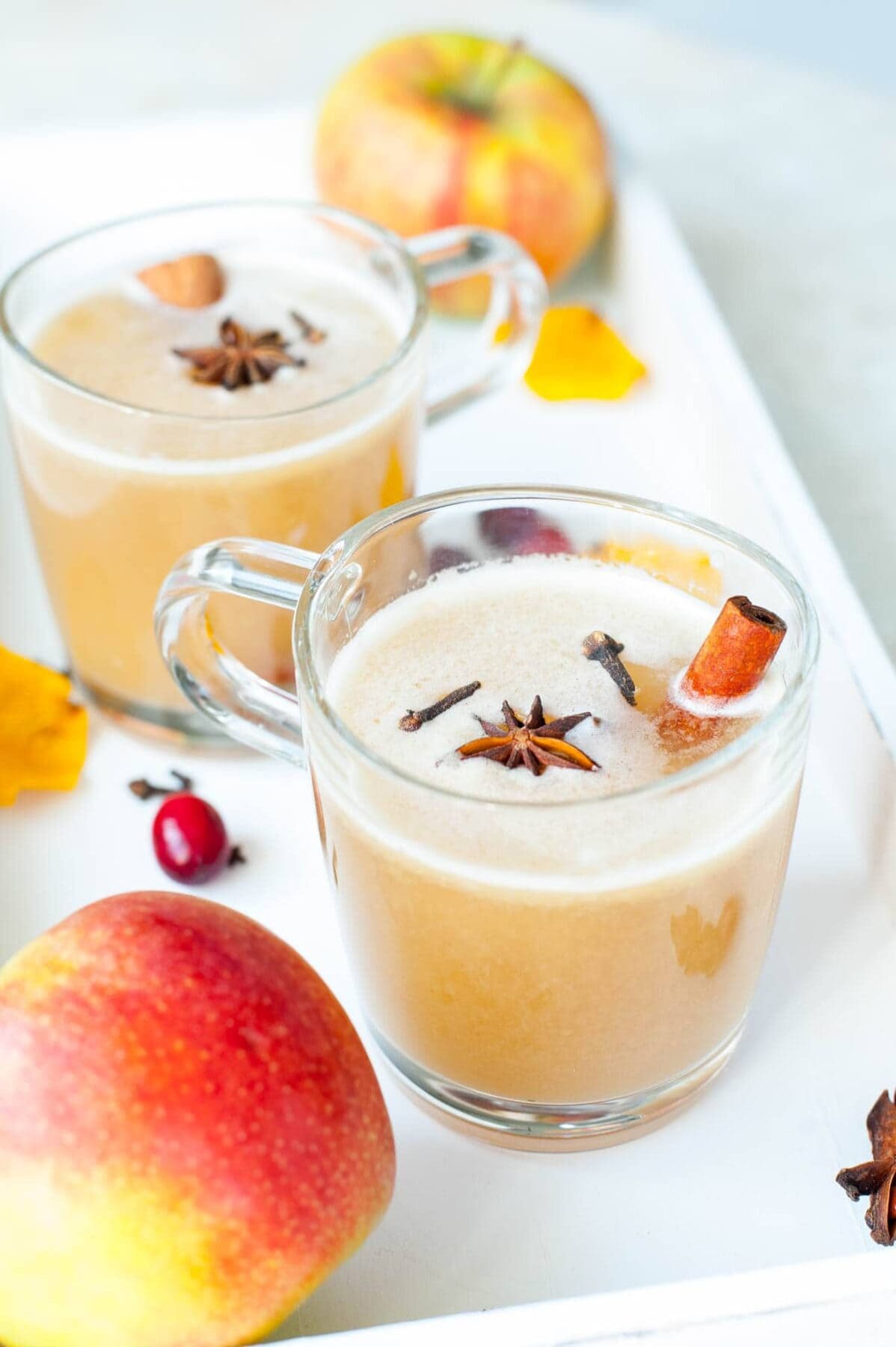 Two glasses with spiced apple cider, apples and spices in the background.