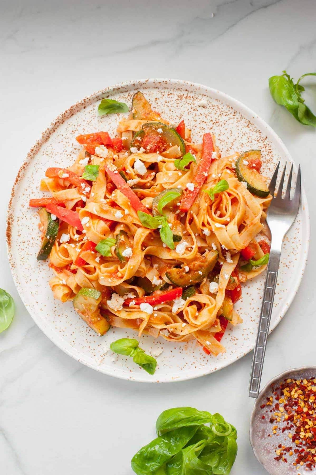 Pasta with zucchini, red bell pepper, tomatoes, and feta cheese on a white plate