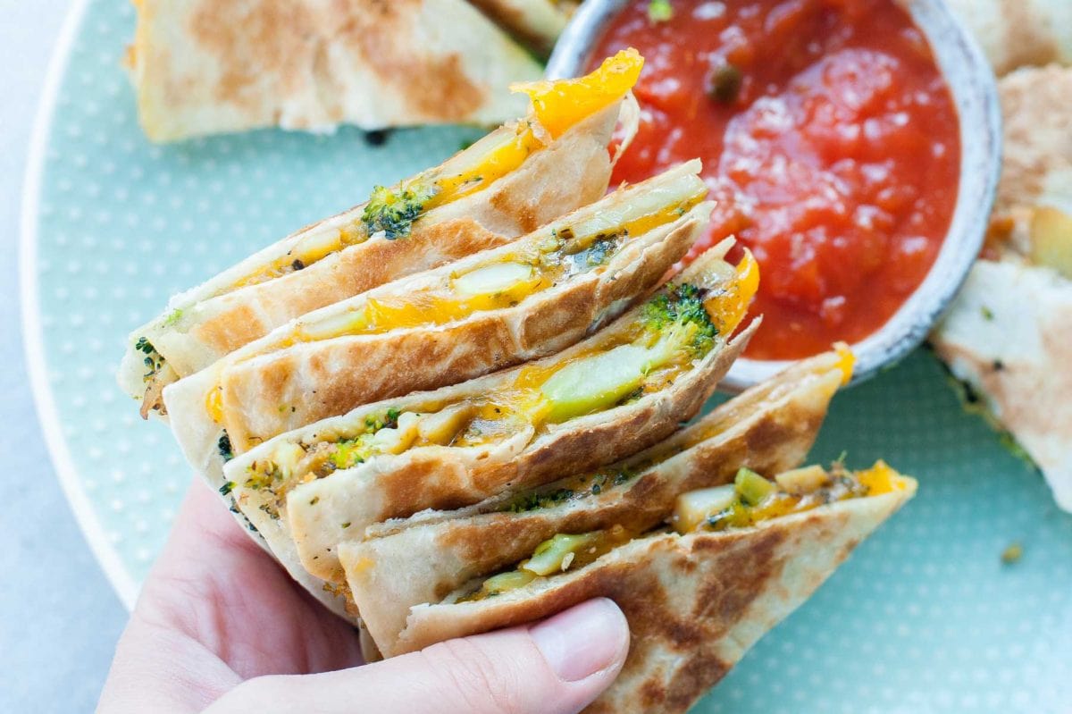 Broccoli cheddar quesadillas are being held in a hand, tomato salsa in the background.