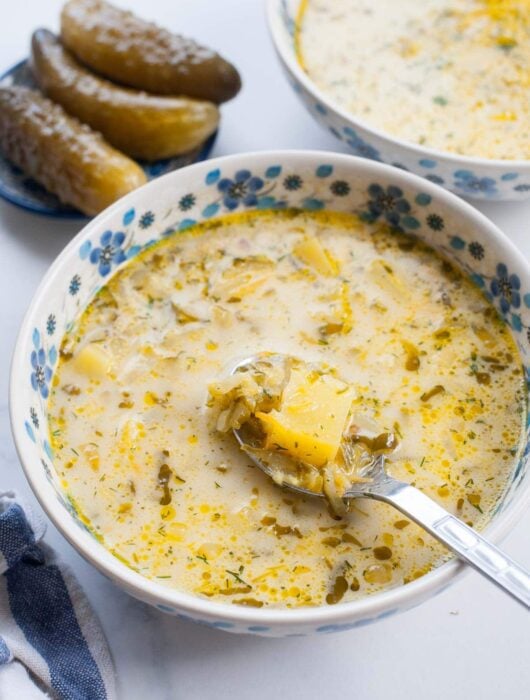 dill pickle soup in a white-blue bowl, fermented cucumbers in the background