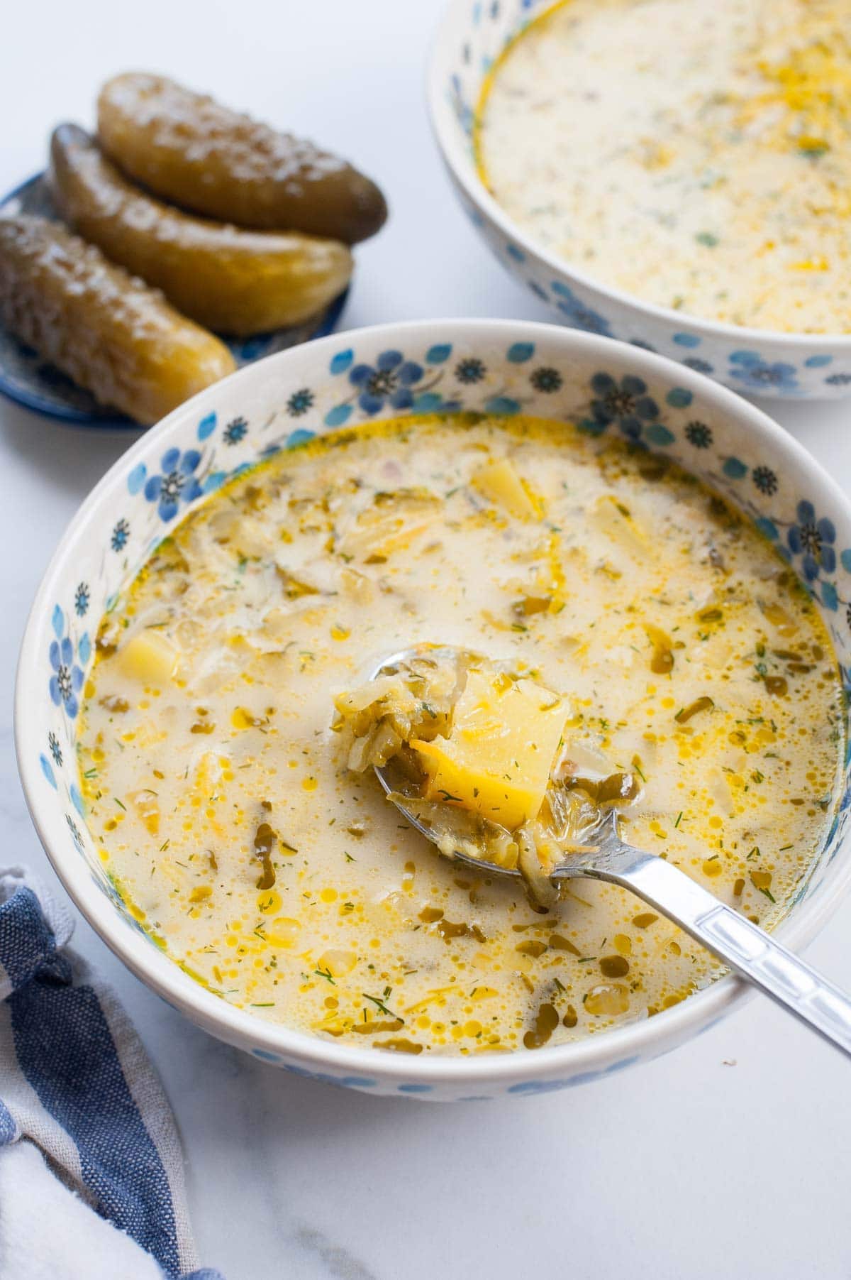 Dill pickle soup in a white bowl with blue flowers, cucumbers in brine on the side.