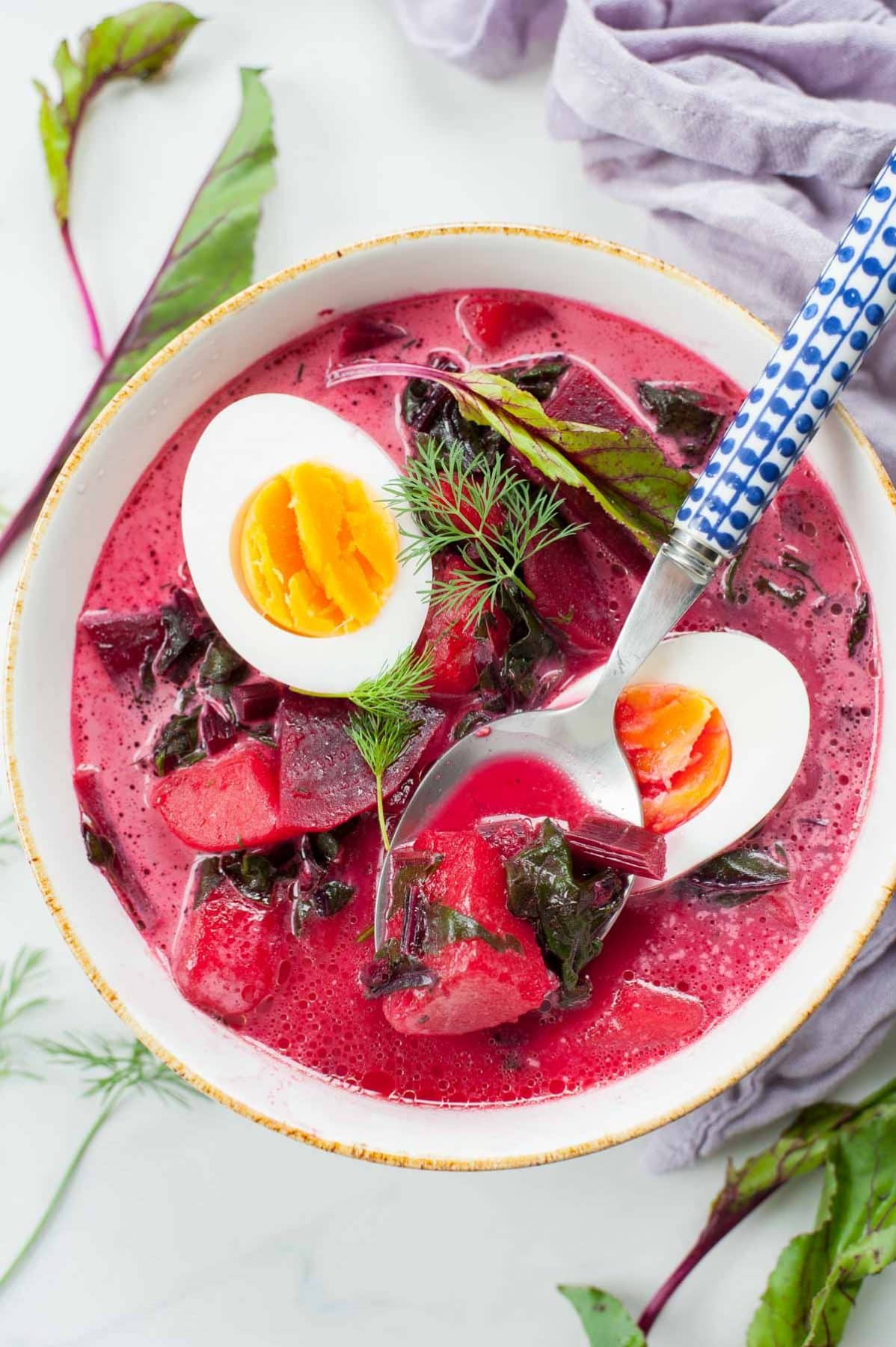 white bowl with beet soup, hard-boiled eggs and dill, blue spoon in the bowl