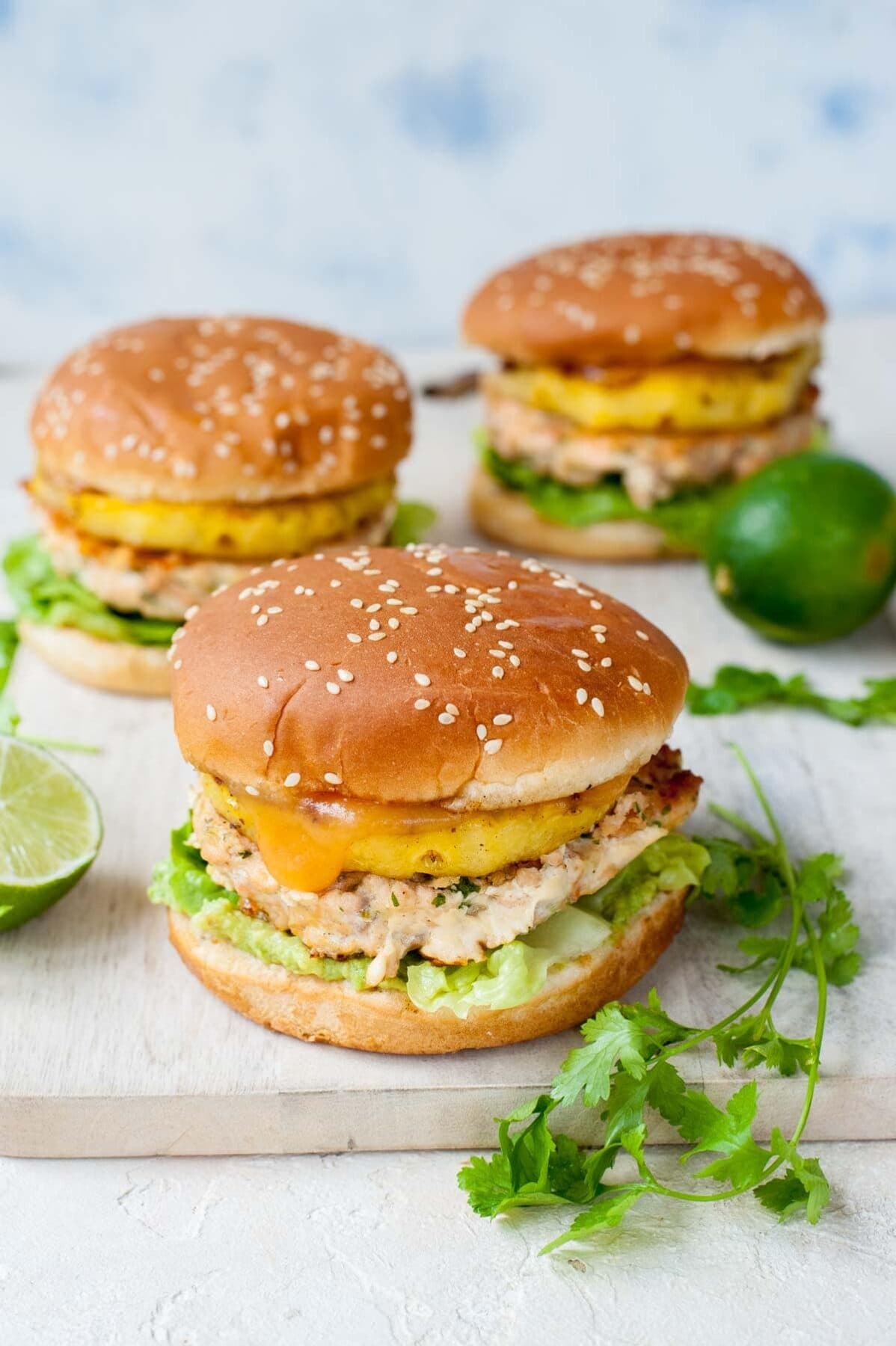 Three salmon burgers with pineapple and guacamole on a beige wooden chopping board.