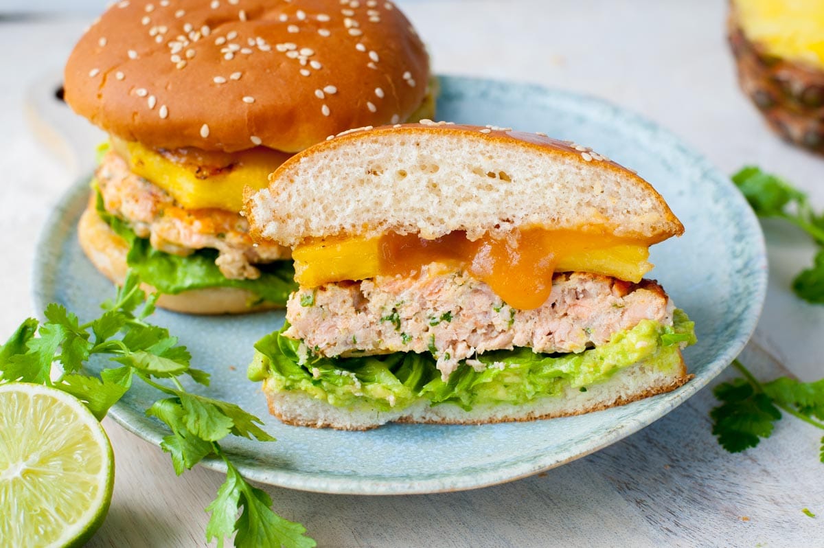 Two salmon burgers on a blue plate, cut in half, cilantro leaves and limes in the background.