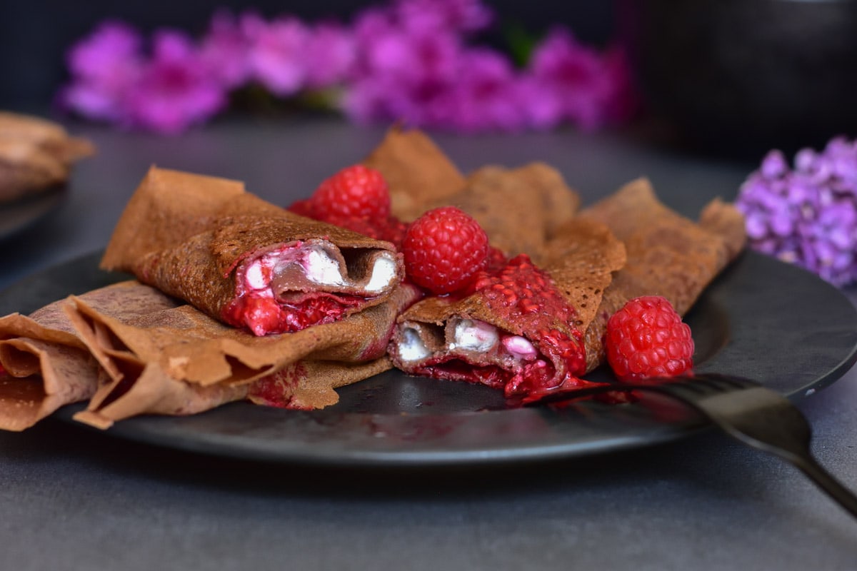 chocolate crepes filled with vanilla cottage cheese and raspberry sauce on a black plate