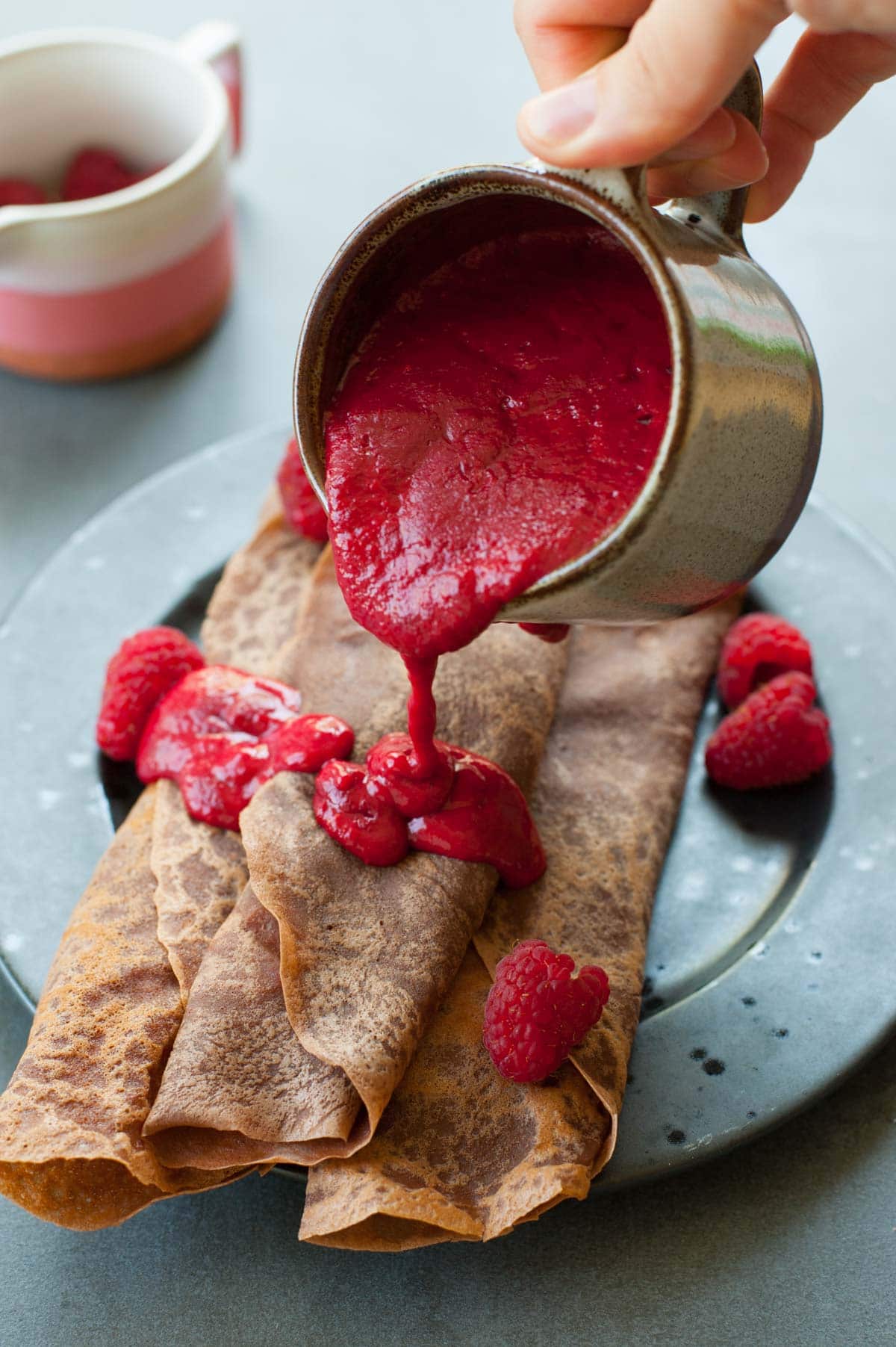 Chocolate crepes on a black plate are being poured with raspberry sauce.