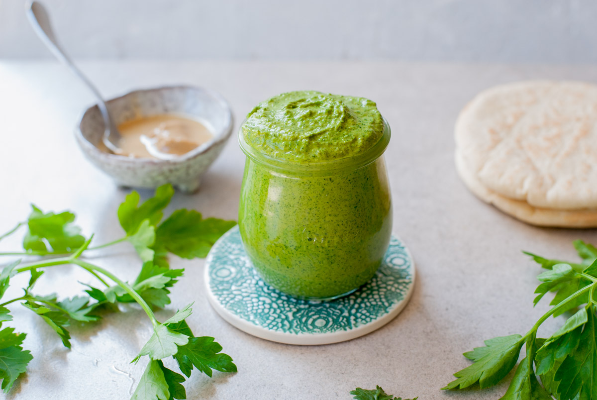 green tahini sauce in a jar, pita bread, tahini paste and parsley leaves in the background