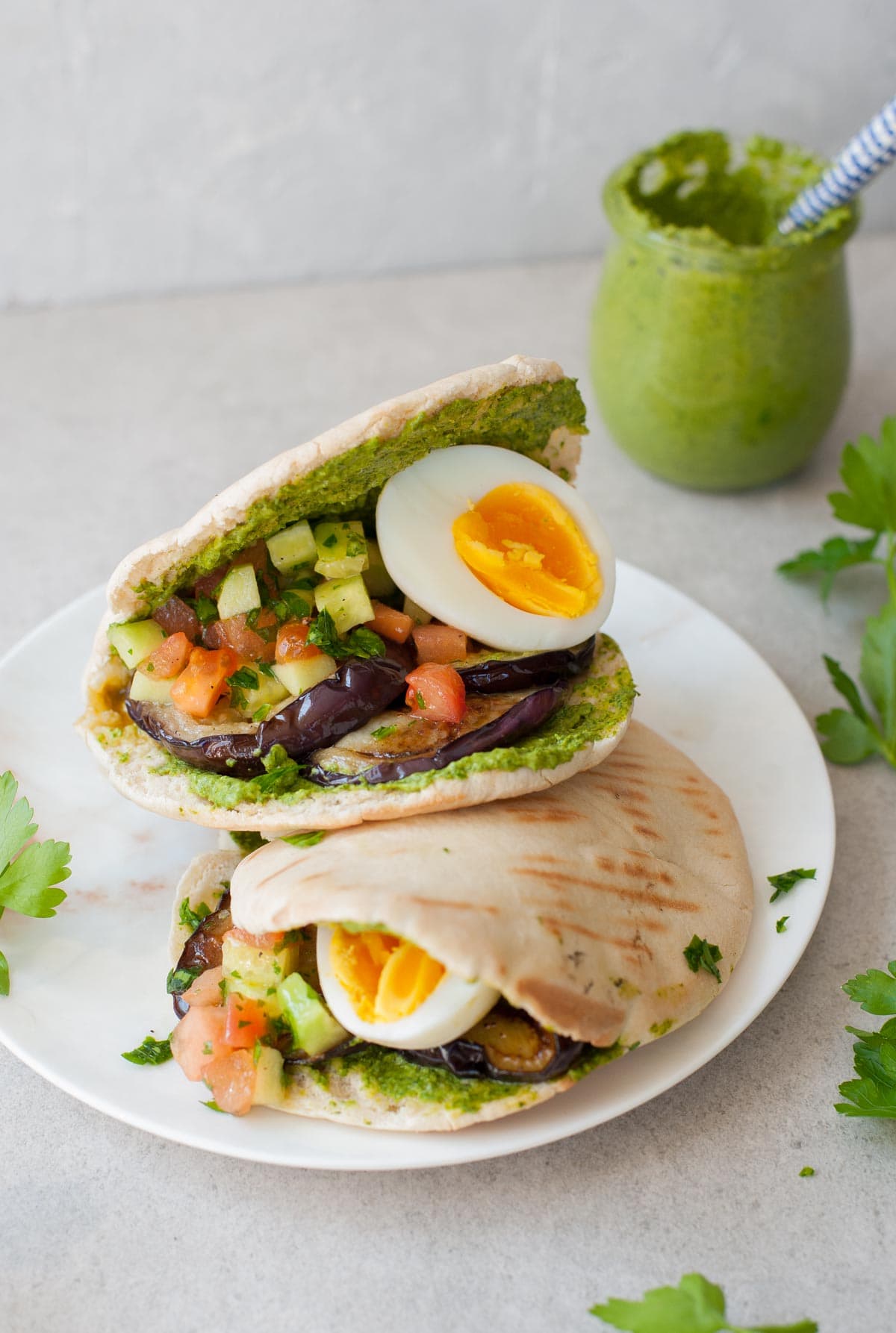 two pita breads filled with eggplant, eggs, tomato and cucumber salad, and green tahini sauce on a white plate