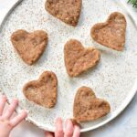 heart-shaped soft gingerbread cookies for babies on a white plate, baby hands reaching for cookie
