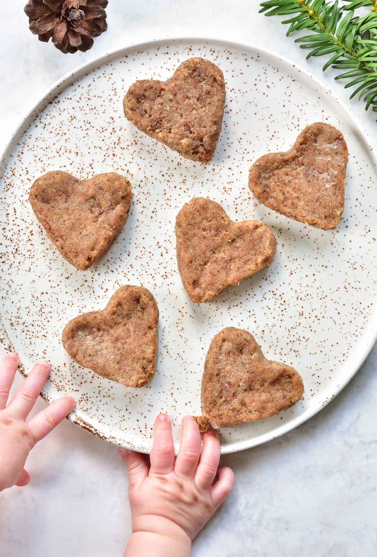 heart-shaped soft gingerbread cookies for babies on a white plate, baby hands reaching for cookie