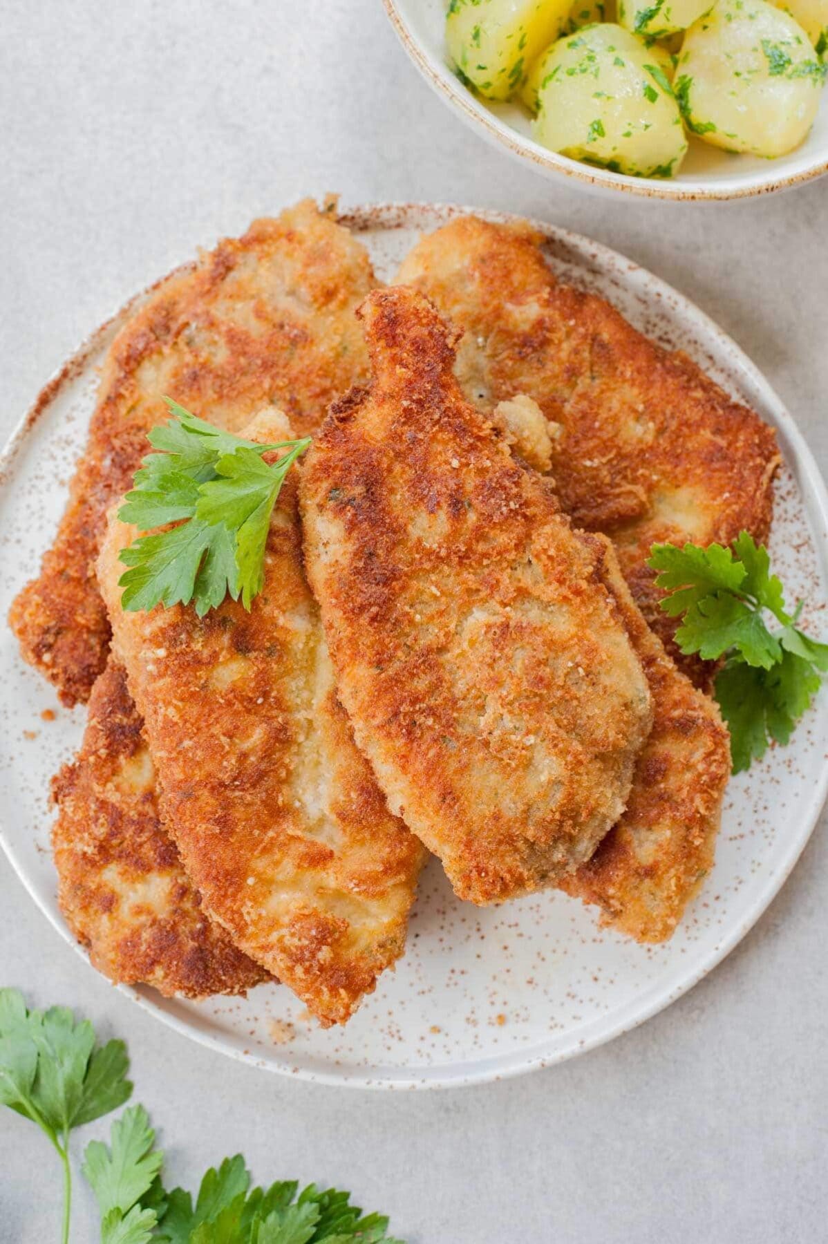 Breaded chicken cutlets on a white plate, parsley potatoes in the background.