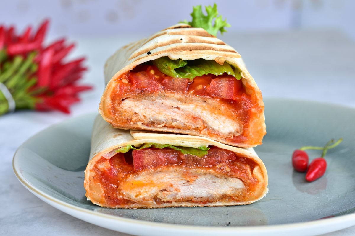 Crispy chicken wrap with spicy tomato salsa cut in half on a blue plate, chili in the background.