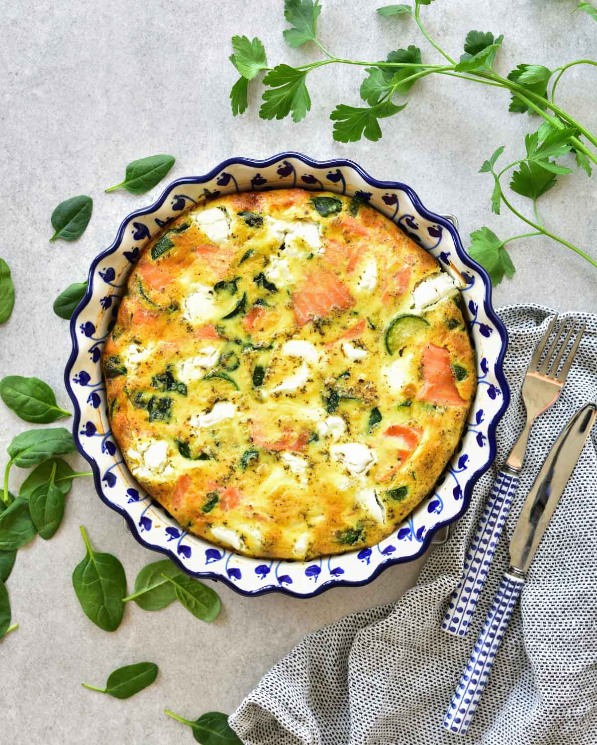 Smoked salmon frittata in a blue baking dish, parsley and spinach in the background.