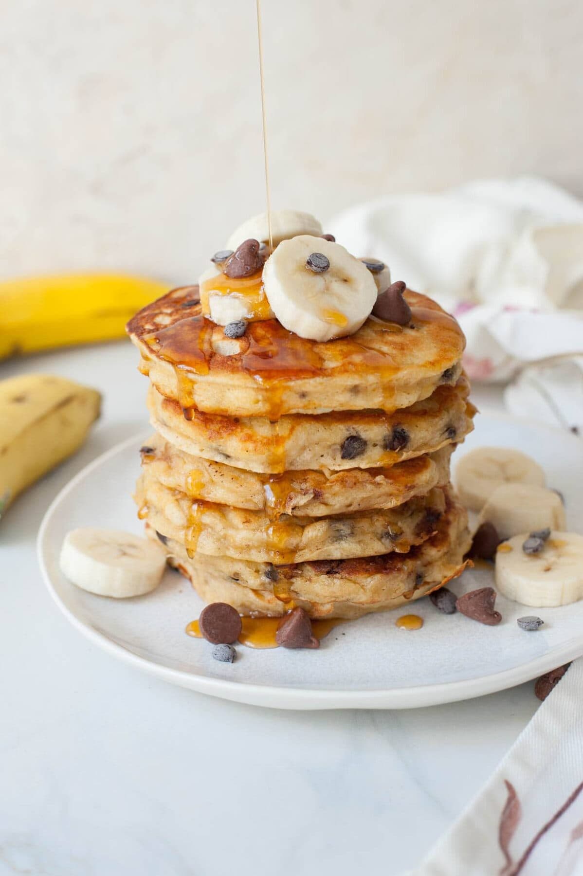 maple syrup is being poured over a stack of banana chocolate chip pancakes