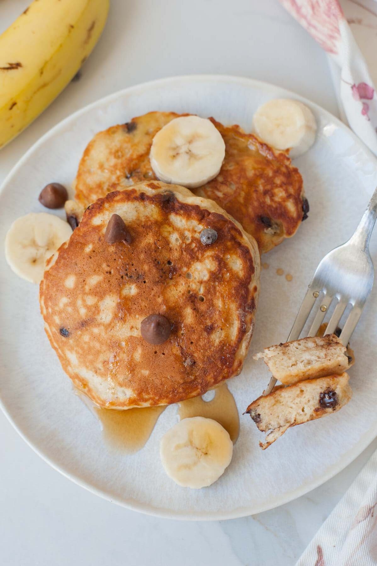 two banana chocolate chip pancakes on a white plate, fork and sliced bananas on the side