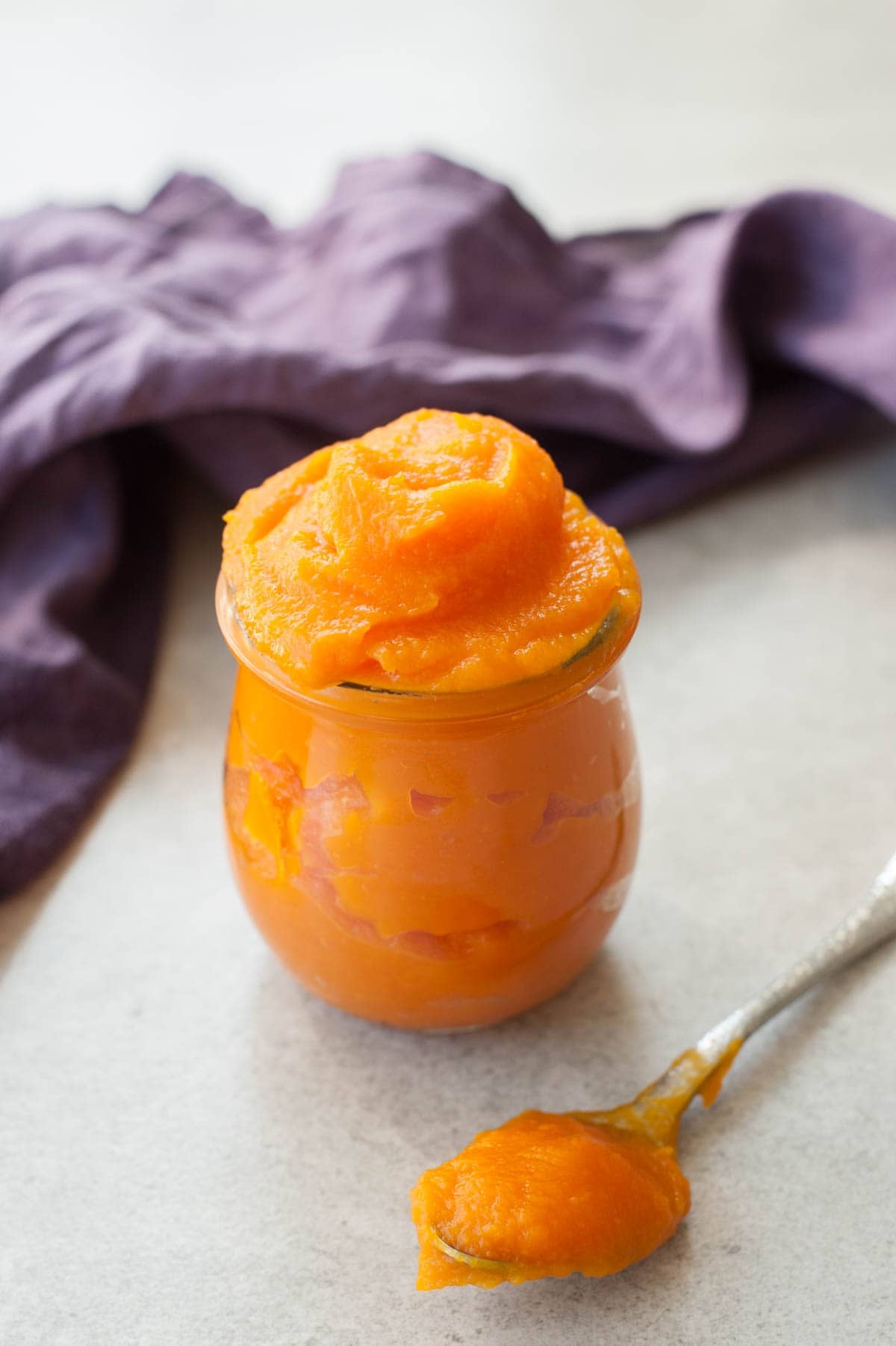 Pumpkin puree in a glass, a spoon with puree in the foreground and a kitchen cloth in the background.