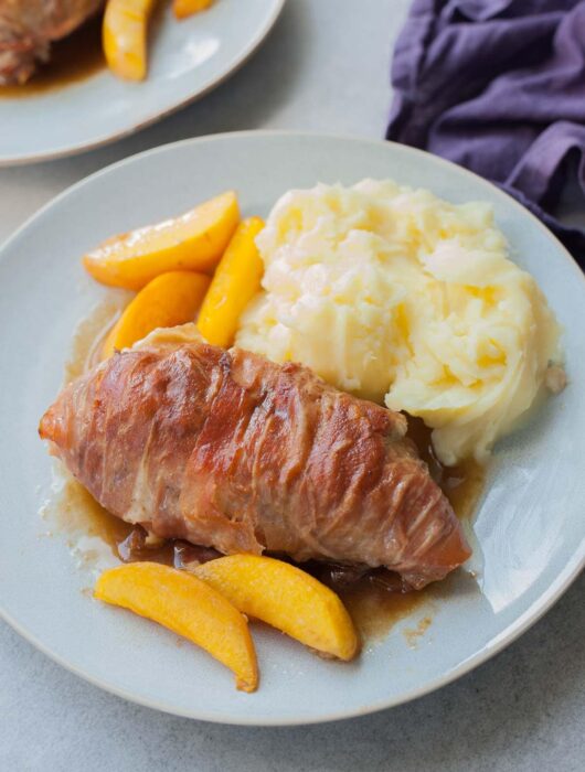 Prosciutto-wrapped chicken with peaches and mashed potatoes on a blue plate.