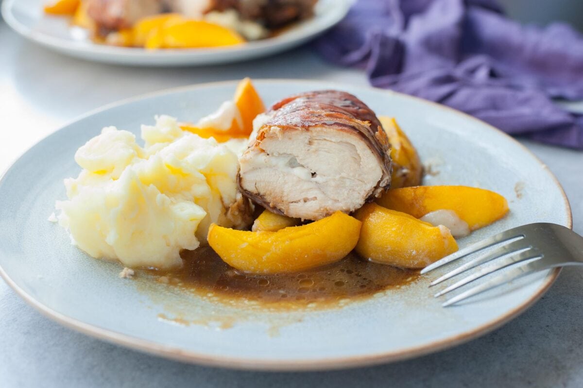 Prosciutto-wrapped chicken cut in half with peaches and mashed potatoes on a blue plate.