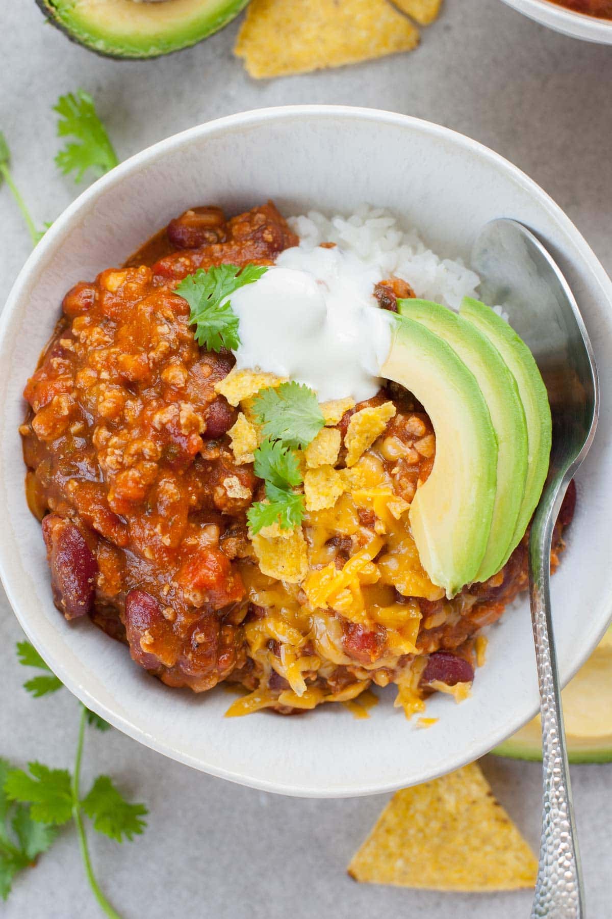 Turkey pumpkin chili with cheddar, avocado and yogurt in a white bowl. Tortilla chips and coriander in the background.