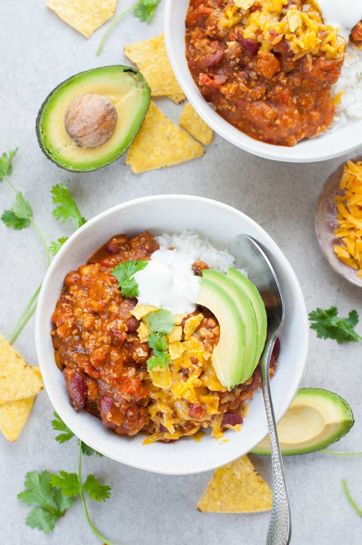Two white bowls with turkey pumpkin chili with cheddar, avocado and yogurt. Tortilla chips and coriander in the background.