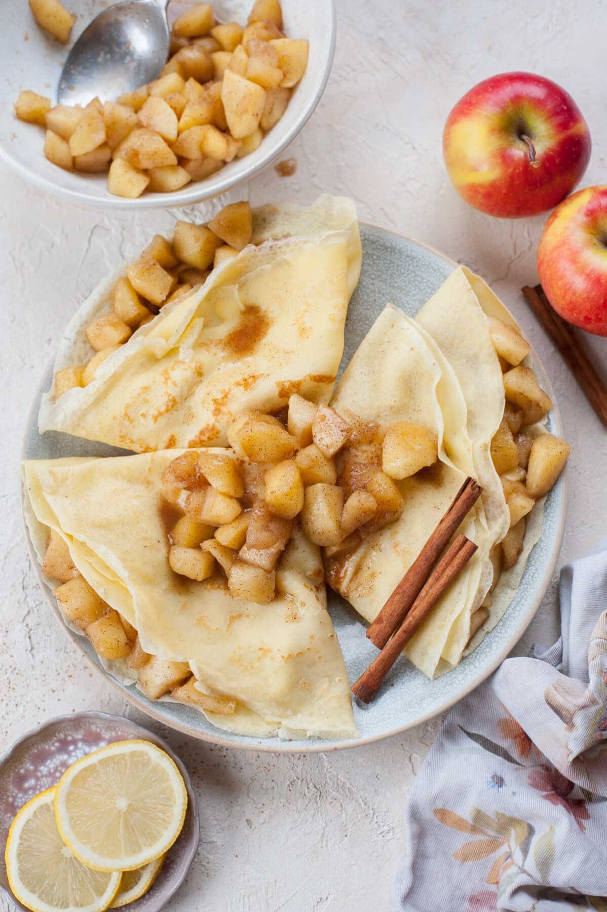 Apple crepes on a blue plate, topped with sauteed apples and cinnamon sticks.