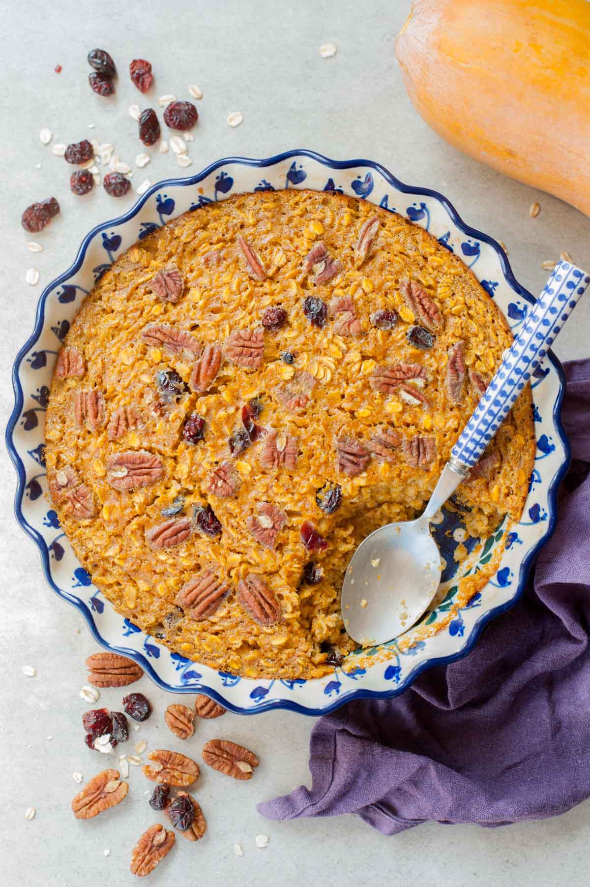 Baked pumpkin oatmeal in a blue-white baking dish. Kitchen cloth, pumpkin, pecans and cranberries in the background.