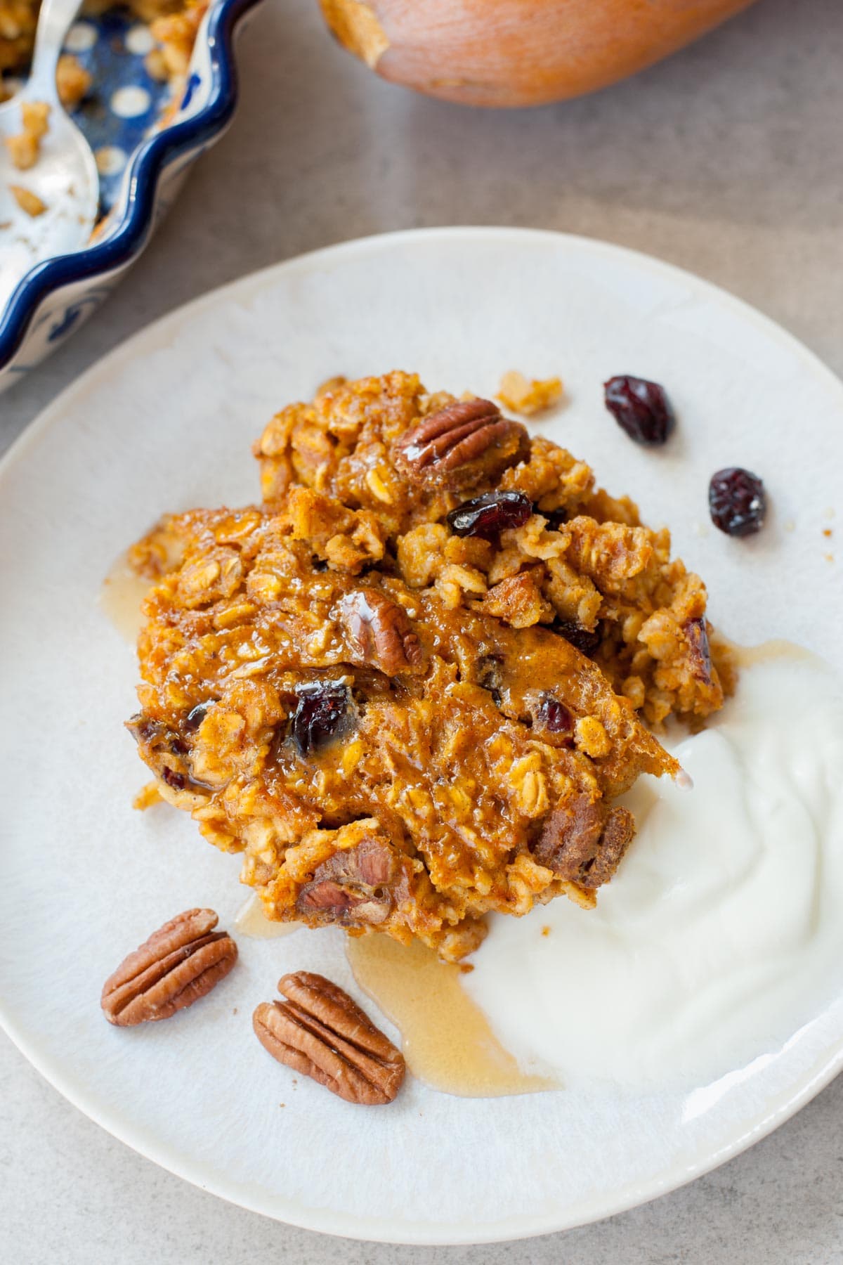 One serving of baked pumpkin oatmeal on a white plate. Baking dish and a pumpkin in the background.