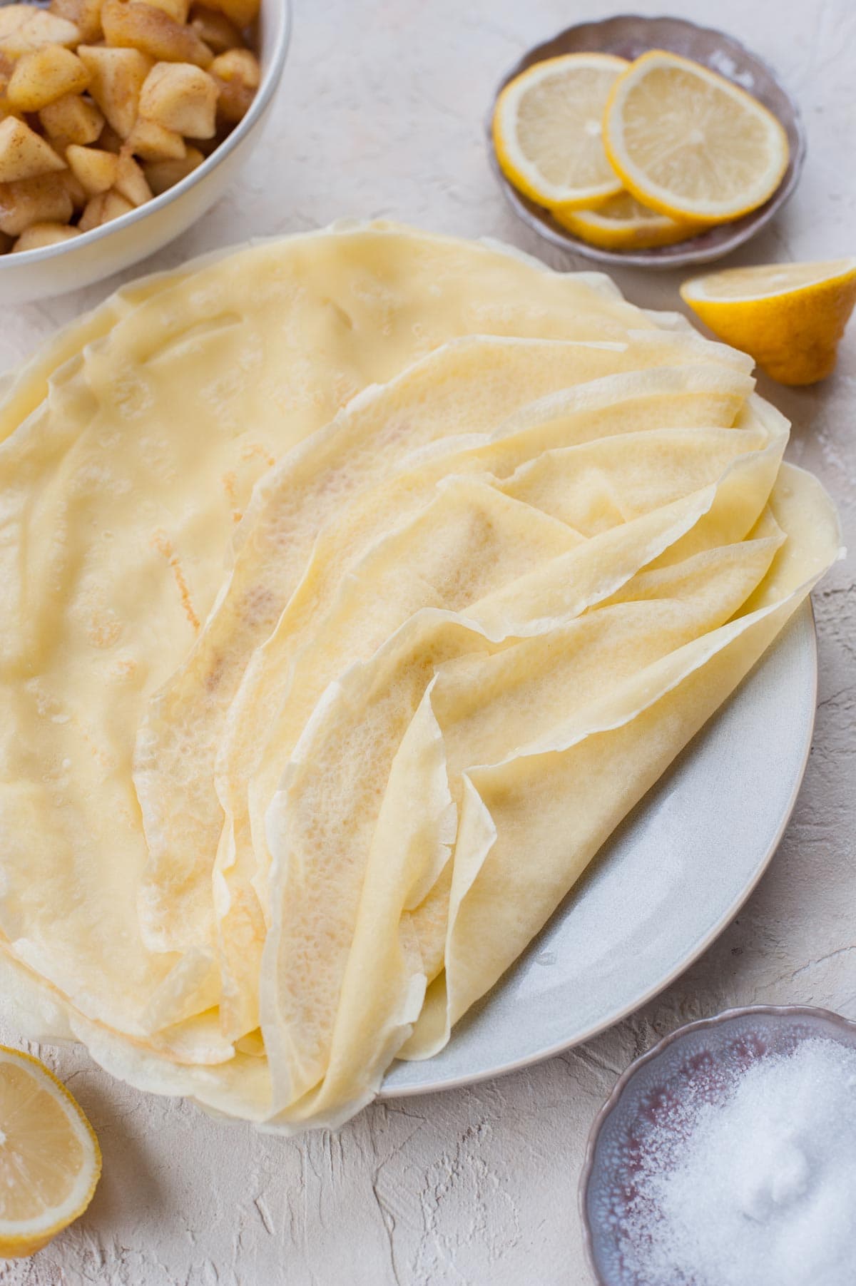 A close up photo of a stack of crepes on a blue plate. Lemons, sugar, and sauteed apples in the background.