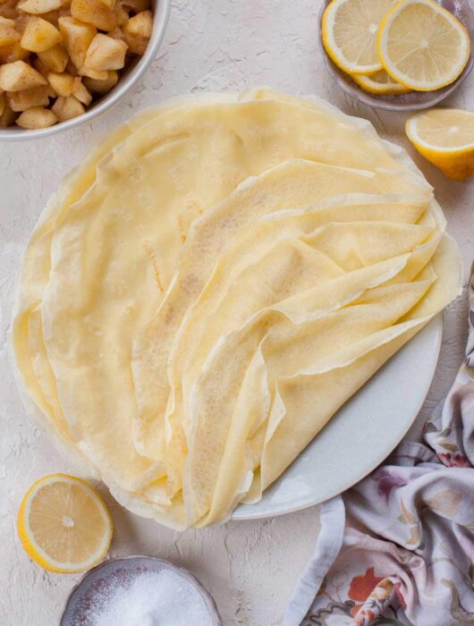 Stack of crepes on a blue plate. Lemons, apples, and sugar in the background.