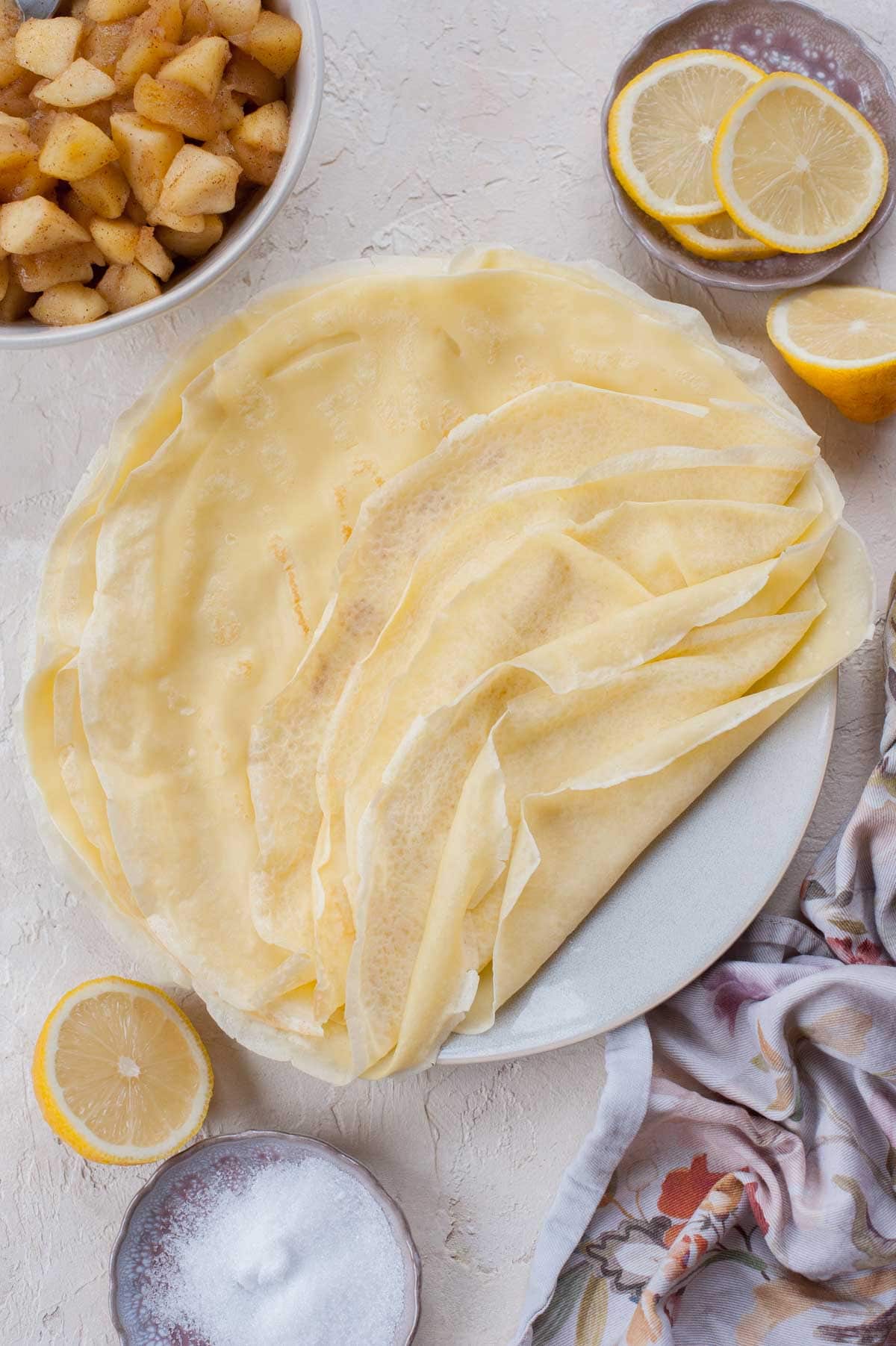 A stack of crepes on a blue plate. Lemons, sugar, and sauteed apples in the background.