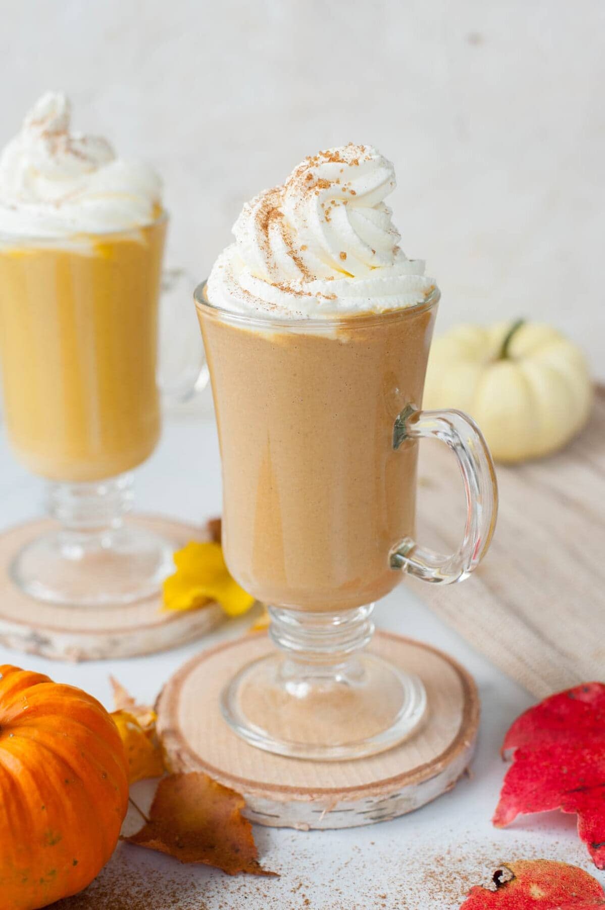 Two glasses with pumpkin hot chocolate topped with whipped cream. Small pumpkin and leaves scattered around.